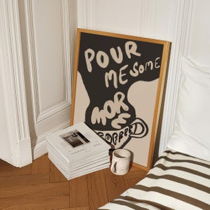 May include: A framed poster with a black background and beige text that reads "Pour me some more coffee". The text is stylized to look like a coffee cup.
