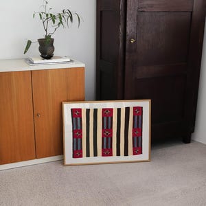 May include: A framed textile art piece with a geometric pattern of vertical stripes in black, beige, and red. The artwork is displayed on a light-colored floor, leaning against a wall near a wooden cabinet and a dark brown wardrobe.