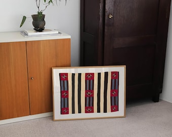 May include: A rectangular framed print featuring a repeating pattern of vertical stripes in cream, black, and blue, interspersed with three wider red bands containing small, dark, cross-shaped geometric motifs centered within the composition.