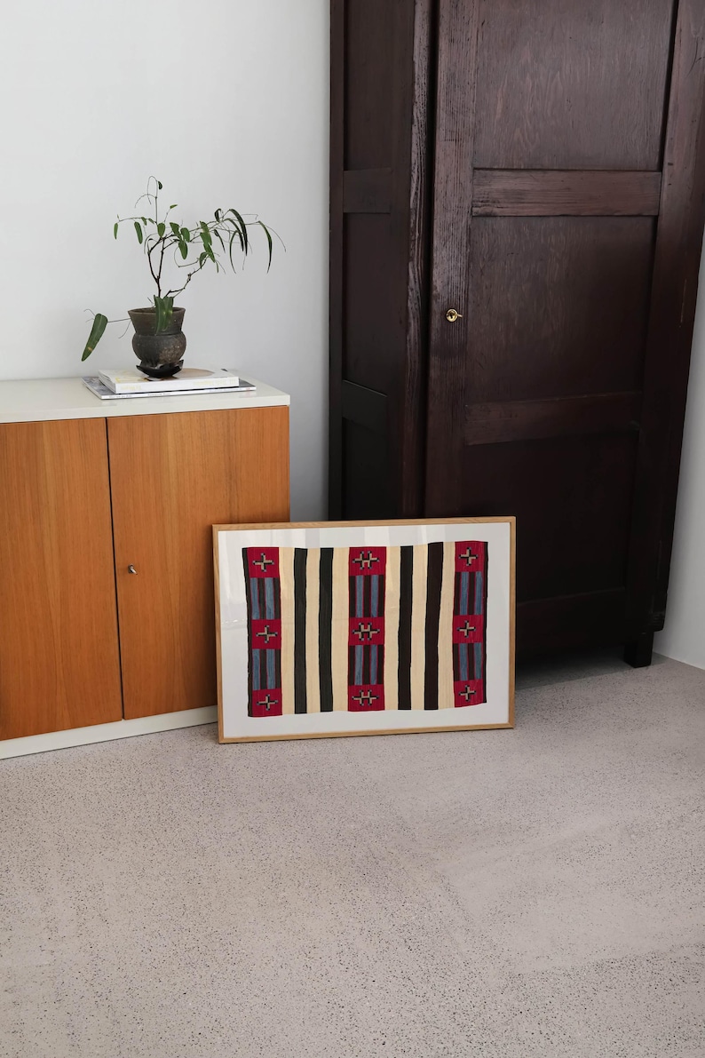 May include: A framed textile art piece with a geometric pattern of vertical stripes in black, beige, and red. The artwork is displayed on a light-colored floor, leaning against a wall near a wooden cabinet and a dark brown wardrobe.