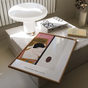 May include: A framed print of a painting by Japanese artist, Mir&oacute; Onchi Koshiro (Japanese, 1891-1955), depicts a nude figure in a pink and gold setting. The print is on a white table with a white lamp and two books, one titled "TOM FORD 002" and the other titled "GENTLE LIVING".