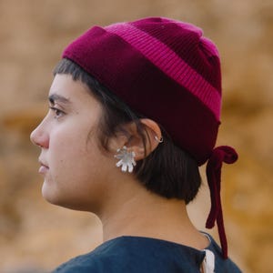 May include: A burgundy and pink striped knit beanie with a matching tie detail. The hat is made of a soft, textured material. The beanie is a stylish accessory for cooler weather.