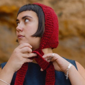 May include: A knitted red bonnet with long ties. The bonnet is made of a textured yarn and is being tied. The background is a blurred natural setting.