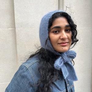 Puede incluir: Una mujer con un gorro y una bufanda de punto azul claro. El gorro está tirado sobre sus orejas y la bufanda está atada en un lazo en su cuello. Lleva una chaqueta vaquera azul oscuro.