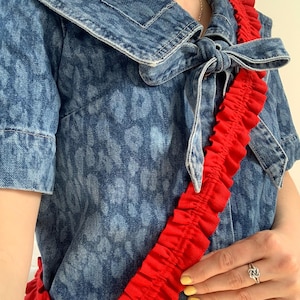 Puede incluir: Una camisa vaquera con estampado de leopardo y un cuello grande atado en forma de lazo. Una correa roja con volantes se lleva sobre la camisa.