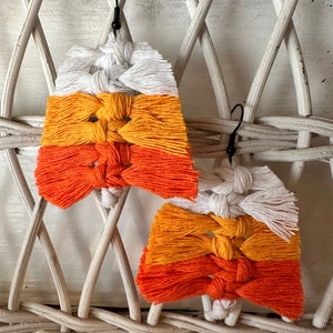 Puede incluir: Un par de pendientes de maíz dulce hechos a mano con hilo blanco, naranja y amarillo. Los pendientes cuelgan de un fondo de mimbre blanco.