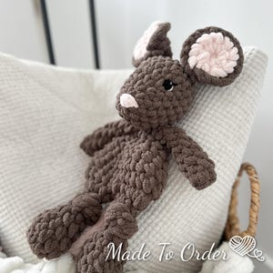 Puede incluir: Un peluche de ratón de ganchillo marrón hecho a mano con detalles rosas en las orejas y la nariz. El peluche está hecho con hilo grueso y está sobre una superficie blanca texturizada. El texto "Made To Order" es visible.