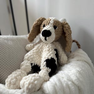 Könnte beinhalten: Handgefertigtes Häkelhund-Plüschtier in Creme, Braun und Schwarz. Der Hund hat Schlappohren, eine schwarze Nase und schwarze Flecken auf seinem Körper. Das Plüschtier liegt auf einer weißen, flauschigen Decke.