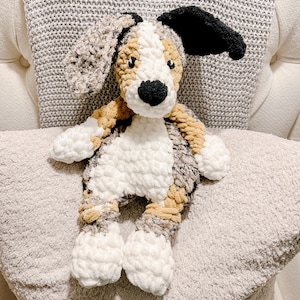 May include: A crocheted dog toy with a black nose, black and white ears, and a white body. The dog has tan and grey accents and is resting on a grey cushion. The toy is soft and cuddly.