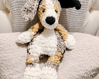 Peut inclure: Une peluche chien au crochet avec un nez noir, des oreilles noires et blanches et un corps blanc. Le chien a des accents beiges et gris et est posé sur un coussin gris. La peluche est douce et câline.