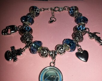 May include: A silver charm bracelet with blue and white charms, including a Carolina Panthers logo charm, a football charm, a heart charm, and a small figure charm. The bracelet is on a pink surface.