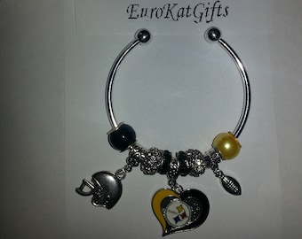 May include: A silver charm bracelet with a Pittsburgh Steelers themed charm. The charm features a heart with the Steelers logo, a football, and a helmet. The bracelet also has black and clear beads.
