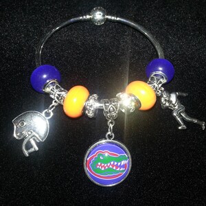Könnte beinhalten: Ein silbernes Armband mit orangefarbenen, blauen und silbernen Perlen. Das Armband hat einen Anhänger in Form eines Footballhelms, einen Anhänger in Form eines Alligators und einen Anhänger in Form eines Footballspielers.
