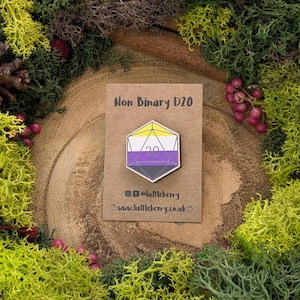 May include: A wooden D20 dice with a non-binary pride flag design. The dice is yellow, white, purple, and black. The dice is on a brown card with the text "Non Binary D20" and a website address.