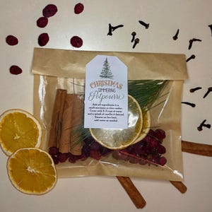Può includere: Un sacchetto di cellophane trasparente pieno di ingredienti per pot-pourri, tra cui bastoncini di cannella, fette d'arancia essiccate e mirtilli rossi. Un'etichetta recita "Christmas Simmering Potpourri". Chiodi di garofano e bacche secche sono sparsi.