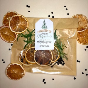 Pode incluir: Um saco de papel castanho cheio de rodelas de laranja secas, raminhos de alecrim e grãos de pimenta, com uma etiqueta que diz "Christmas Simmering Potpourri". Rodelas de laranja secas adicionais são dispostas ao redor. Uma fragrância festiva para casa.