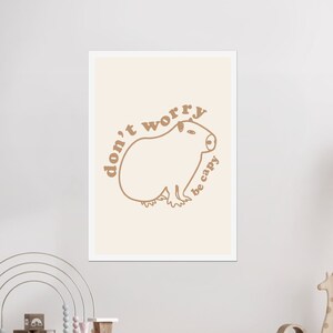 Pode incluir: Uma impressão emoldurada bege com um contorno marrom de um capivara e o texto "don't worry be happy" em marrom.