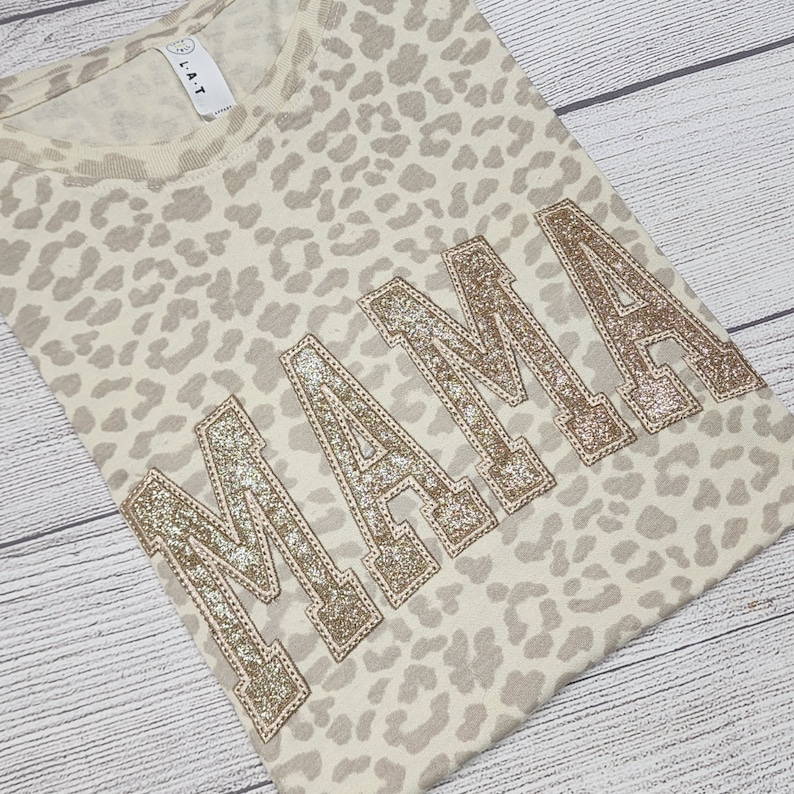 May include: A beige t-shirt with a leopard print pattern and the word "MAMA" in gold glitter lettering.