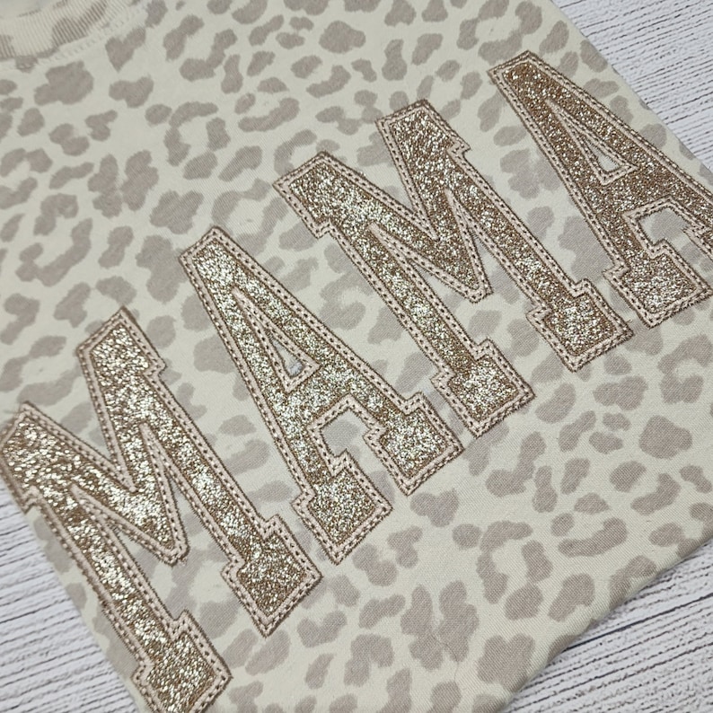 May include: A beige sweatshirt with a leopard print pattern and the word "MAMA" in gold glitter lettering.
