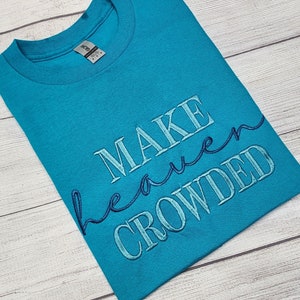 Puede incluir: Camiseta de cuello redondo color turquesa con la frase bordada "MAKE heaven CROWDED" en hilo azul claro y azul marino. La camiseta está doblada sobre una superficie de madera clara.