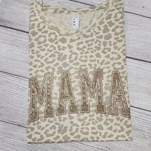 May include: A beige t-shirt with a leopard print pattern. The word "MAMA" is written in large, gold glitter letters across the front of the shirt.