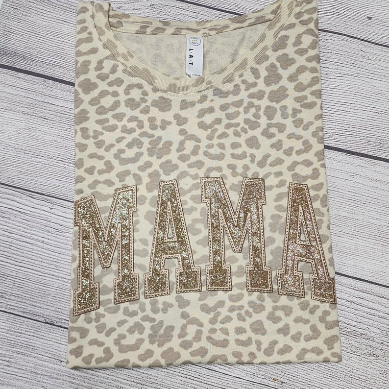 May include: A beige t-shirt with a leopard print pattern. The word "MAMA" is written in large, gold glitter letters across the front of the shirt.