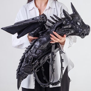 May include: A woman is holding a black leather dragon-shaped bag with a detailed design. The bag has a long strap and a large head with a toothy grin.