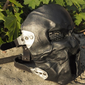 Op de afbeelding: Een zwart lederen masker met een gevormde gezichtsstructuur, met gaas bedekte oogopeningen en een grote open mond. Het masker is bevestigd met zilverkleurige metalen zijplaten, klinknagels en verstelbare zwarte lederen riemen met gespsluitingen.