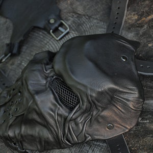 May include: A black leather mask with a mesh covering the eyes and a textured surface. The mask has a strap that goes around the head.