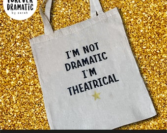 May include: A white tote bag with black text that reads "I'm not dramatic I'm theatrical" and a gold star. The bag has black handles and is on a gold glitter background.