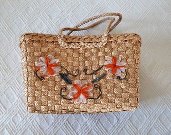May include: A woven straw tote bag with a brown rope handle and orange and white flower embellishments.
