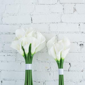 May include: Two bouquets of white calla lilies with green stems, tied with white ribbon. The flowers are set against a white brick wall, creating a clean and elegant aesthetic. Ideal for weddings or home decor.
