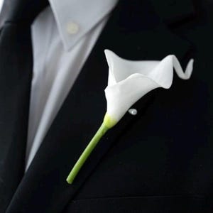 May include: A white calla lily buttonhole pinned to a black suit jacket. The flower has a long green stem and a small pearl pin. The suit is paired with a white dress shirt and a black tie, creating a formal look.