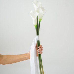 May include: A bouquet of three white calla lilies with green stems and accents. The flowers are tied with a white ribbon. The bouquet is held by a person's hand against a white background.