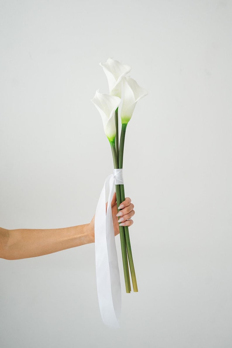 May include: A bouquet of three white calla lilies with green stems and accents. The flowers are tied with a white ribbon. The bouquet is held by a person's hand against a white background.