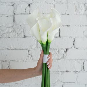 May include: A bouquet of white calla lilies with green stems, held together with a white band. The flowers have a smooth, elegant appearance. The background is a white brick wall.