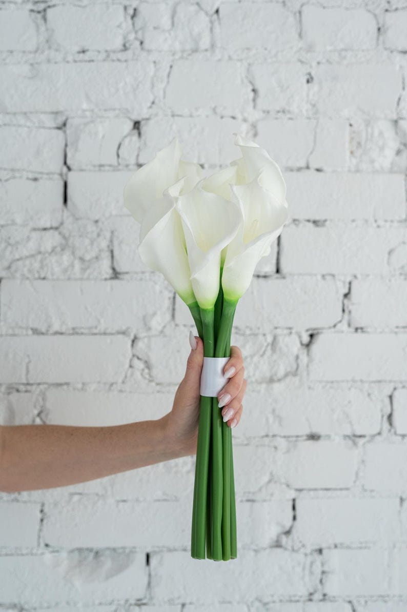 May include: A bouquet of white calla lilies with green stems, held together with a white band. The flowers have a smooth, elegant appearance. The background is a white brick wall.