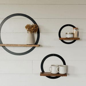 May include: Three circular wall shelves with black metal rings and wooden shelves. One shelf holds a vase with dried flowers, another a candle and matches, and the third two white mugs. The shelves are mounted on a white, wood-paneled wall.
