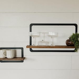 May include: Two black metal shelves with wooden shelves. The top shelf has three clear glasses and a small brown vase with green plants. The bottom shelf has two white mugs.