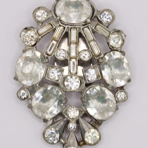 May include: A silver-toned metal brooch featuring a symmetrical arrangement of clear, faceted glass stones. The design includes large oval-cut stones, smaller round-cut stones, and rectangular baguette-cut stones set in a tiered, fan-like pattern.
