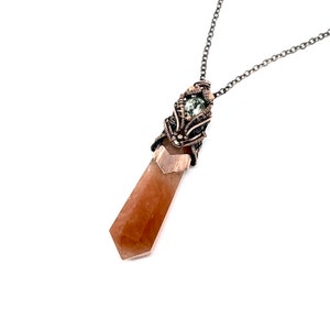 May include: A pendant necklace featuring a faceted, light brown crystal point. The crystal is encased in a decorative, dark metal wire wrap, with a small green stone accent. The necklace chain is a matching dark metal.
