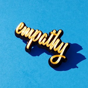 May include: A gold-colored word "empathy" is written in a cursive font on a black background. The word is on a blue surface.