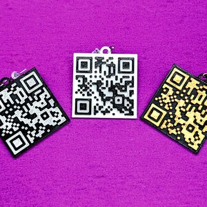 Op de afbeelding: Drie vierkante QR-code oorbellen, een zwart-wit, een wit en een gouden. De oorbellen zijn op een paarse achtergrond.