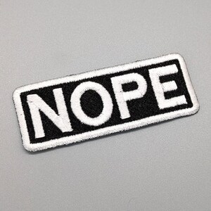 May include: A black rectangular patch with white embroidered text that reads "NOPE".