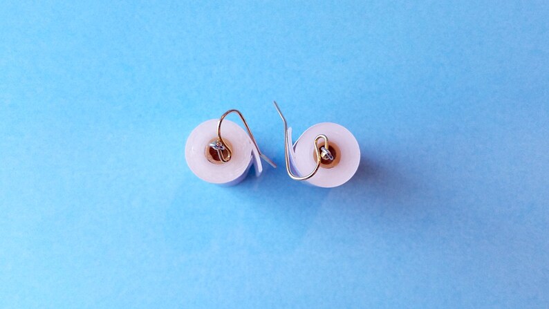 May include: A pair of white toilet paper roll earrings with silver hooks. The earrings are on a blue background.