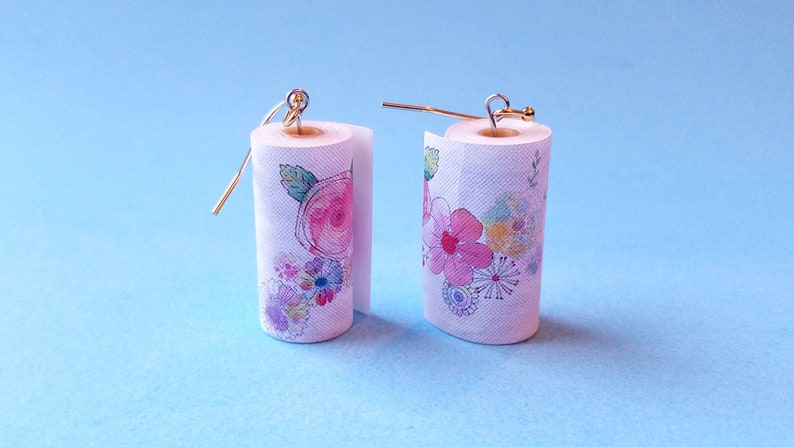 May include: A pair of earrings shaped like paper towel rolls with a floral design. The earrings are white with pink, blue, and green flowers.