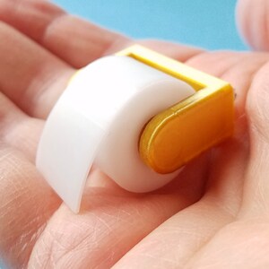 May include: A miniature gold-colored toilet paper holder with a white roll of paper. The holder is small enough to fit in the palm of a hand.