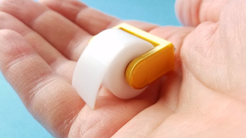 May include: A miniature gold-colored toilet paper holder with a white roll of paper. The holder is small enough to fit in the palm of a hand.