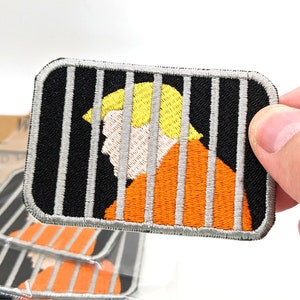 May include: An embroidered iron-on patch featuring a cartoon of a person with blond hair in an orange jumpsuit behind bars. The patch is black, orange, and gray.