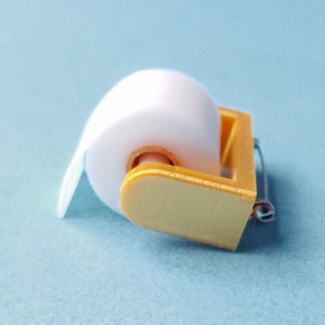 May include: A miniature gold-colored toilet paper holder with a white roll of paper.